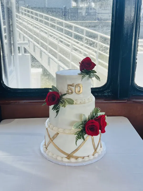 Three-tier white cake with gold criss-cross ribbon, gold 50 numerals and clusters of red roses