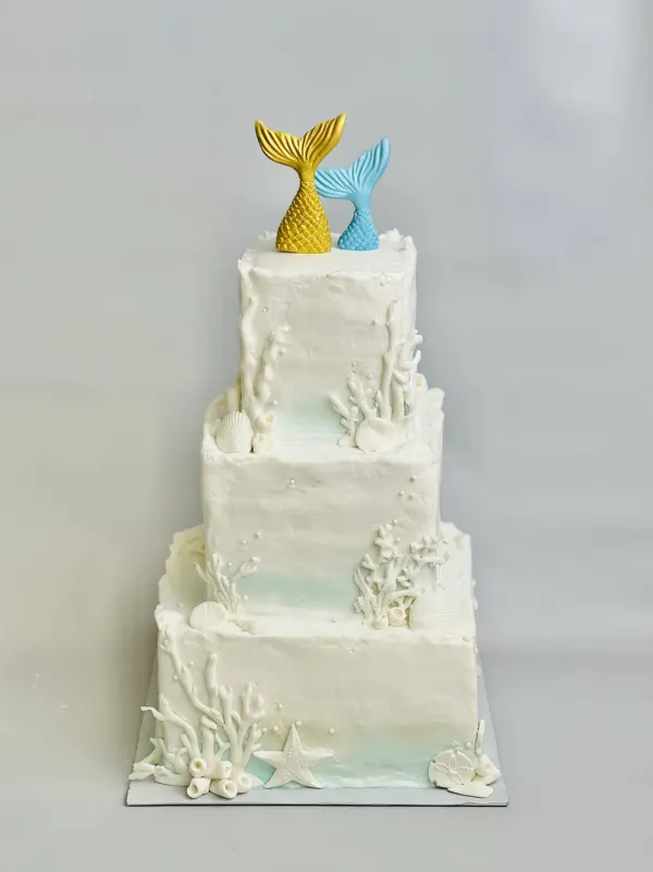 Three-tier square white cake with sugar coral, shells, starfish and gold and blue mermaid tails