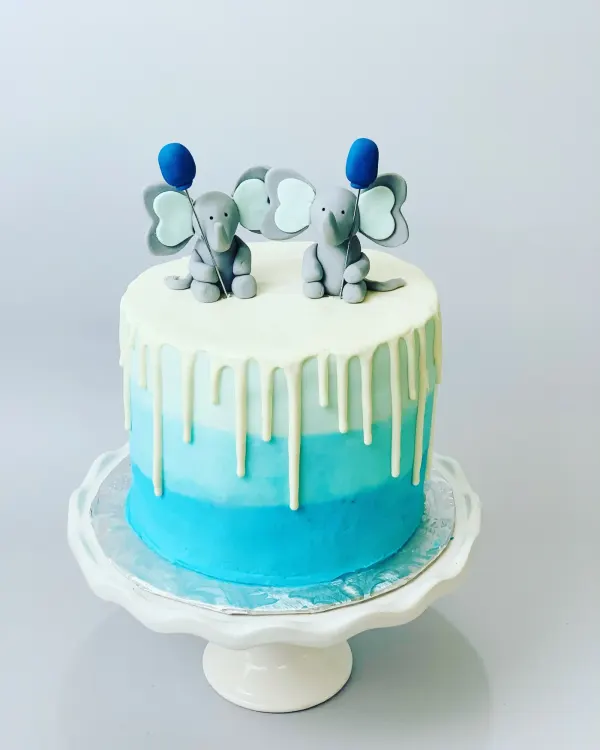 Blue ombre cake with white chocolate drip and two grey fondant elephants holding blue balloons