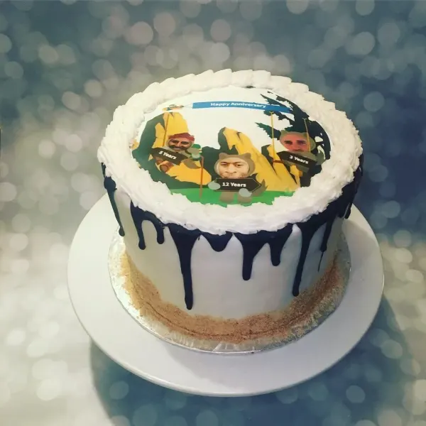 White cake with navy drip and cookie crumb base topped with a Happy Anniversary photo of three hikers