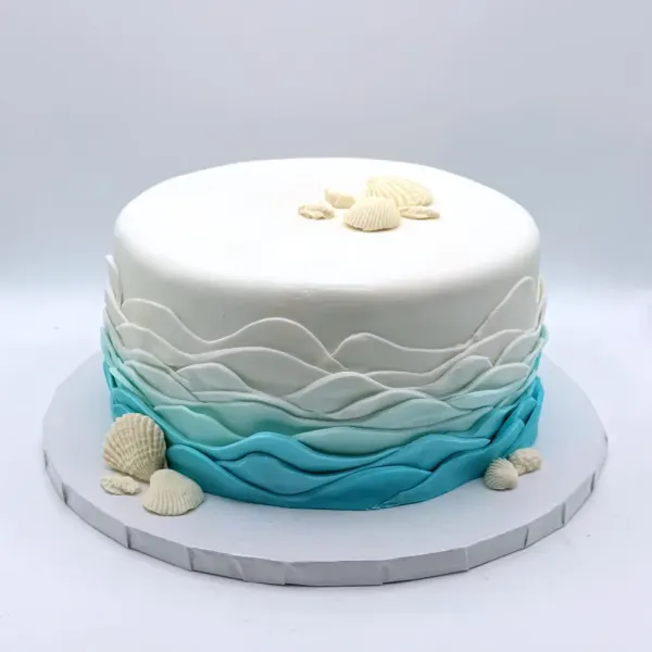 White cake with blue ombré waves around the base and white fondant seashells