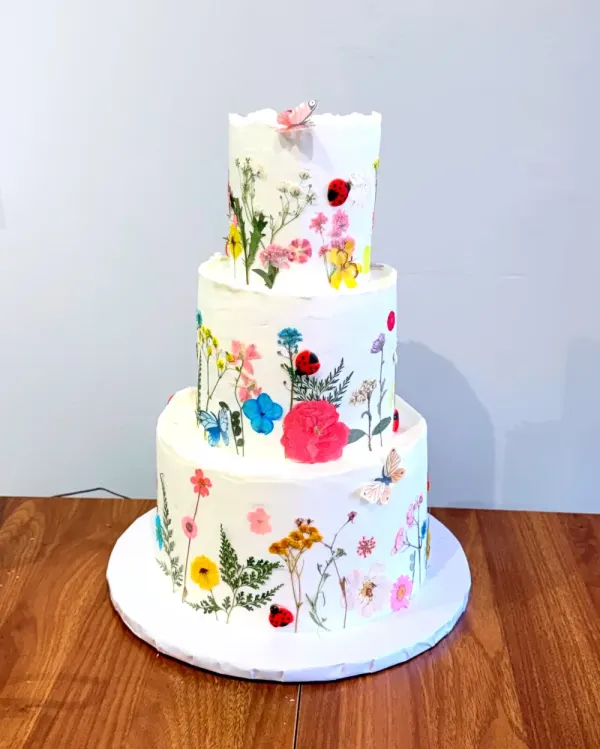 Three tier white cake covered in pressed wildflowers in pink, red, blue and yellow