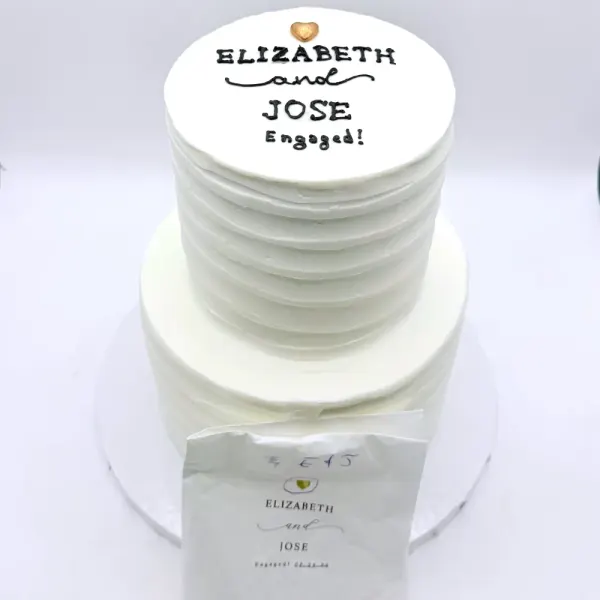 Two tier white cake with a ribbed top tier and a round plaque lettered for Elizabeth and Jose