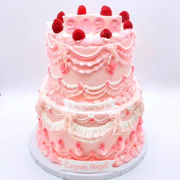 Two tier pink vintage piped cake with ruffles, swags, raspberries on top and Congrats Abigail at the base