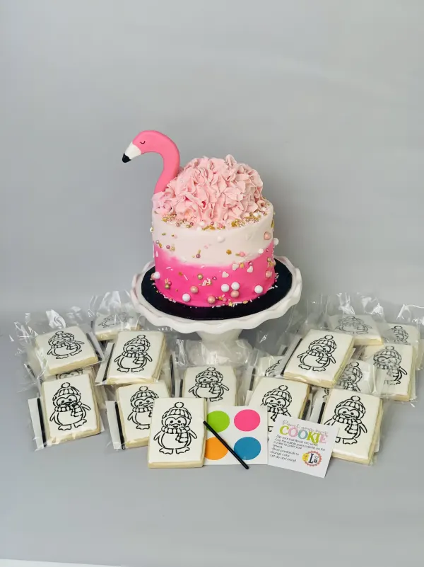 Pink ombre ruffle cake with flamingo topper beside packaged paint-your-own penguin cookies