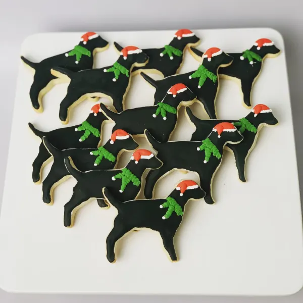 Black Labrador silhouette cookies in Santa hats with piped green holly collars on a white platter