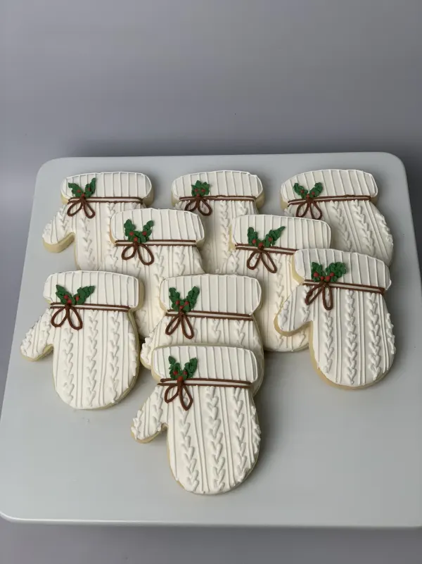 Eight white cable-knit mitten cookies with brown piped bows and green holly sprigs