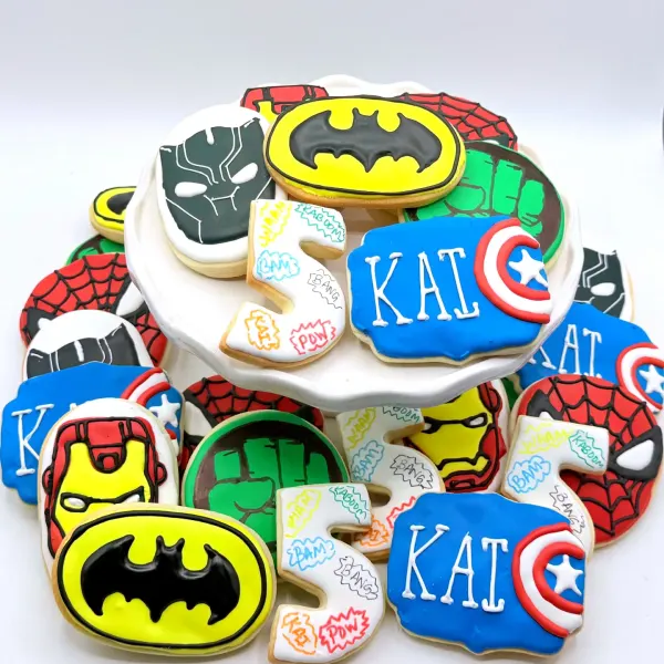 Superhero cookies with masks, shields and emblems in bold colors, with Kai lettered plaques