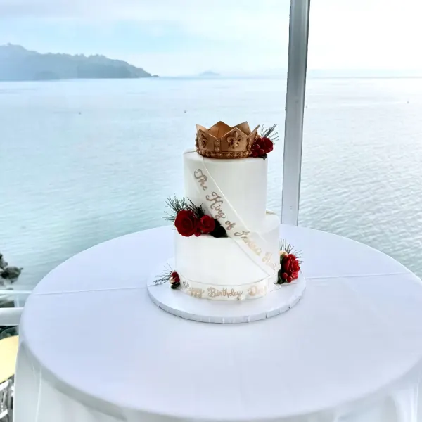 Two tier white cake with a gold crown topper and red berry sprigs, on a table overlooking the water