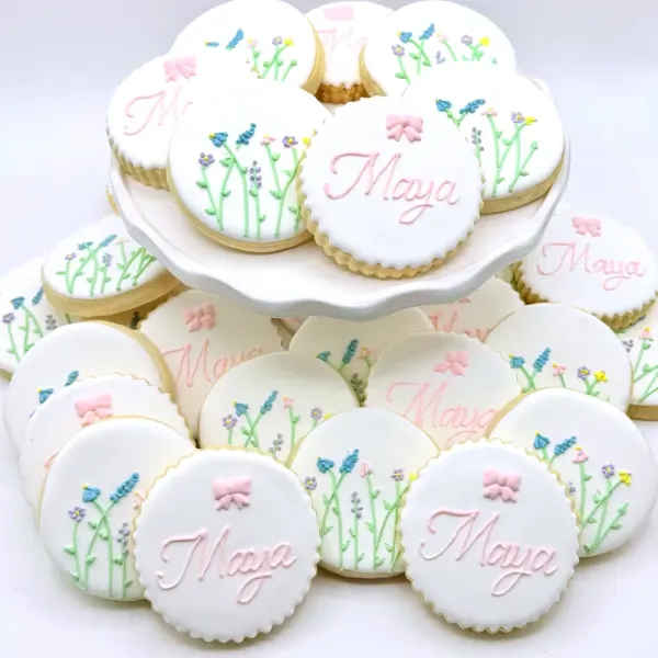 White cookies with pastel wildflower sprigs, pink bows and Maya piped in pink script