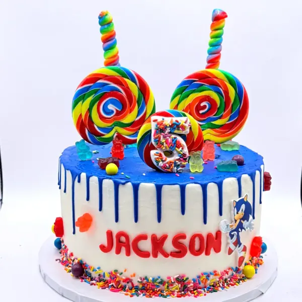 Blue drip cake topped with rainbow lollipops and gummy bears, lettered Jackson, with a sprinkle base