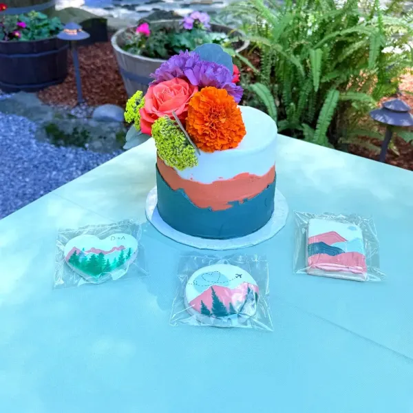 Blue and coral mountain landscape cake with a floral topper beside individually wrapped matching cookies