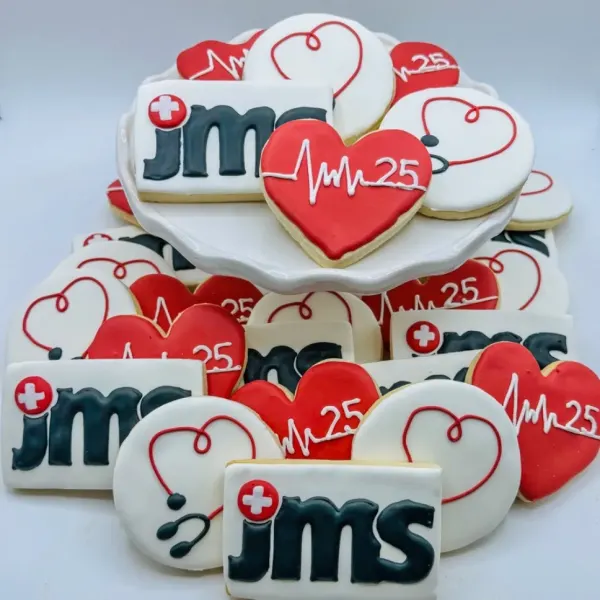 A tiered display of decorated sugar cookies including rectangles with a navy and red jms logo, red hearts piped with a white heartbeat line and the number 25, and round cookies decorated with a red heart and stethoscope design.