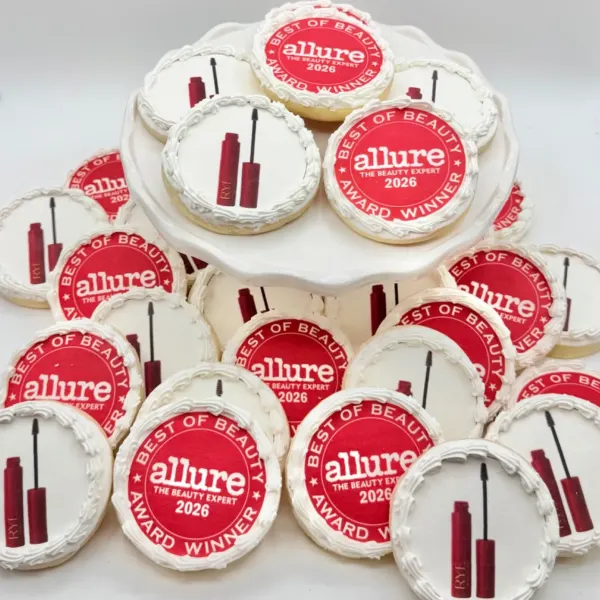 A stack of round frosted cookies alternating between a red and white award seal design and a mascara tube and wand illustration, displayed on a white pedestal stand.