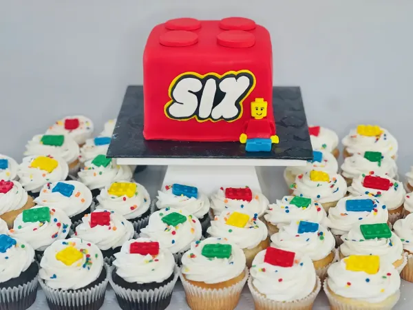 Red building brick cake reading SIX with a minifigure and cupcakes topped with fondant bricks