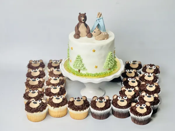 Woodland cake with bear and fawn figures and piped pine trees beside chocolate bear face cupcakes