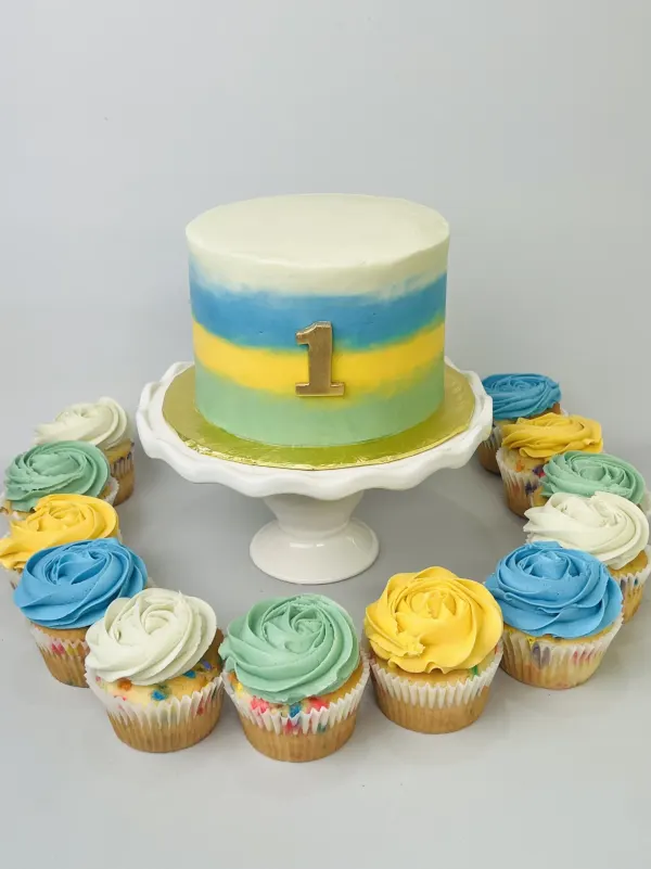 Blue, yellow and green striped cake with a gold number 1 ringed by buttercream rosette cupcakes