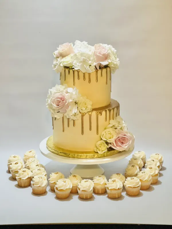 Two-tier gold drip cake with fresh pink and white roses beside mini cupcakes with gold star toppers