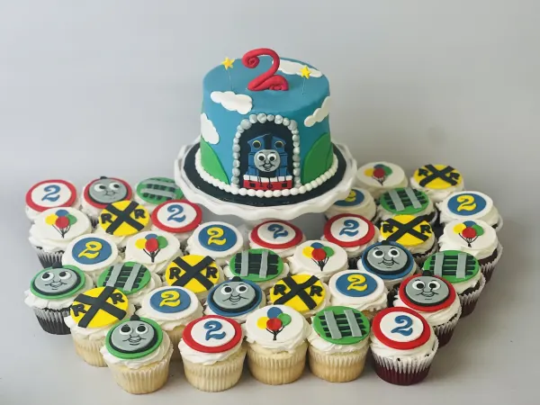 Blue train cake with a fondant number 2 beside cupcakes with train faces and railroad crossings