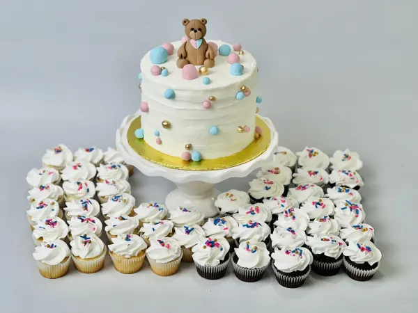 Smooth white cake with a fondant teddy bear and pink, blue and gold dots, ringed by sprinkle cupcakes