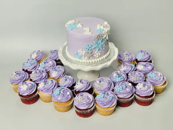 Lavender snowflake cake with a fondant number 1, ringed by lavender cupcakes with snowflake sprinkles