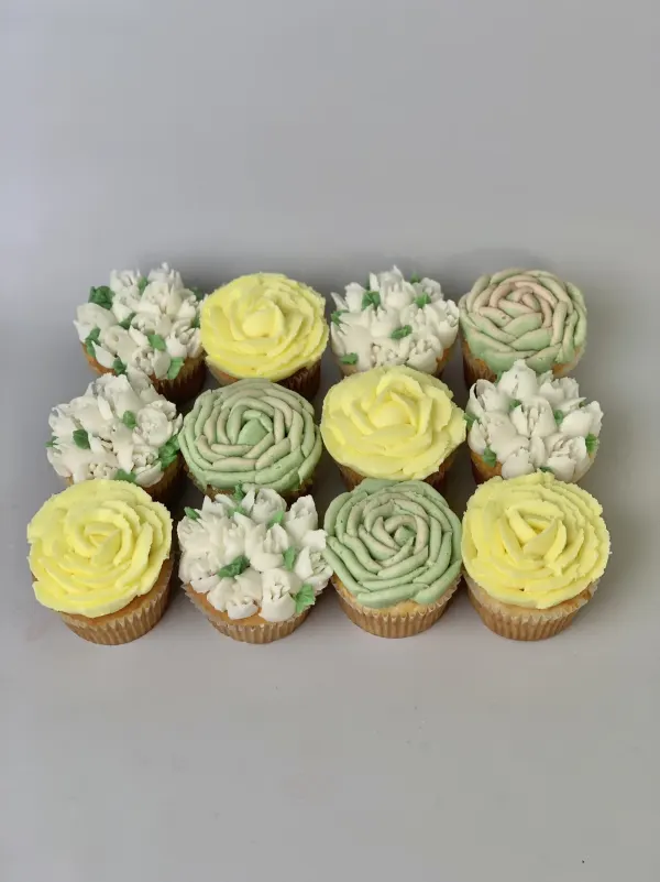 Twelve cupcakes piped with buttercream roses and tulip clusters in yellow, white and sage green