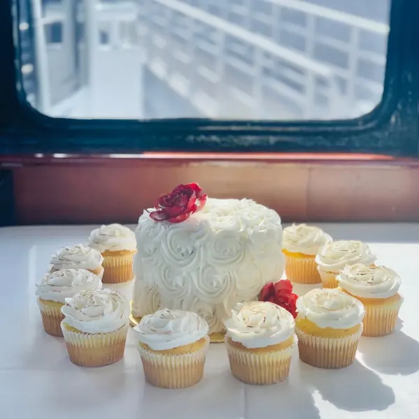 Cake covered in white buttercream rosettes with red blooms, ringed by white swirl cupcakes