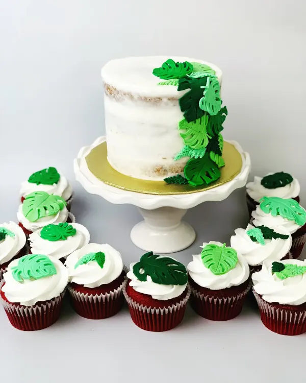 Semi-naked cake trimmed with green fondant monstera leaves, plus red velvet cupcakes with leaf toppers