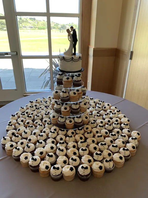 White cake with black piped swags and a bride and groom figurine above cupcakes with black bow ties