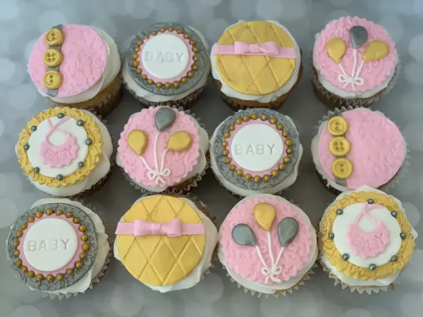 Baby shower cupcakes with pink, gold and silver discs reading BABY, plus bibs, balloons and buttons