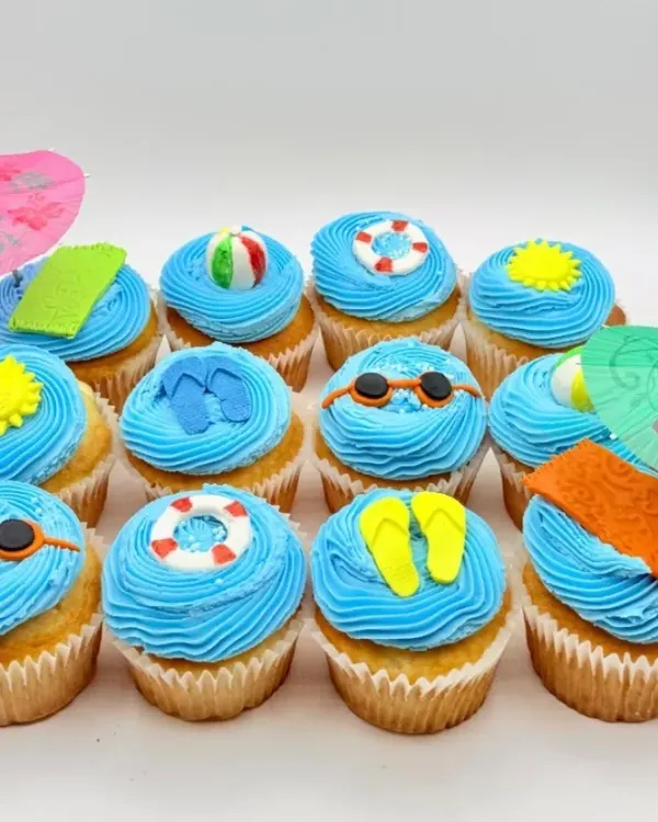 A dozen vanilla cupcakes with blue buttercream swirls topped with fondant sunglasses, flip flops, beach balls, pool floats, and towels, decorated with pink and green paper cocktail umbrellas.