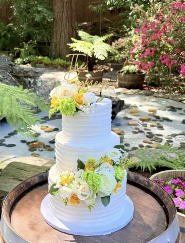 Meghan three-tier wedding cake in horizontally combed white buttercream with yellow and white roses, craspedia and green hydrangea, standing on a wine barrel in a fern garden, 2026