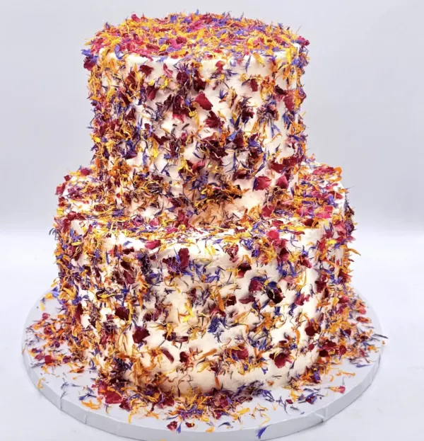 Julia two-tier wedding cake covered entirely in dried edible flower petals in cornflower blue, deep red and marigold, 2026