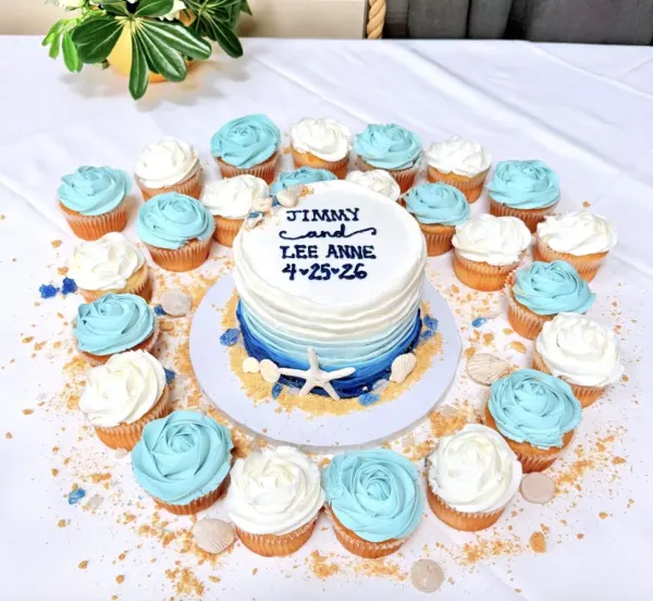 Jimmy and Lee Anne single-tier white wedding cake with a blue ombre base, starfish and sand, lettered Jimmy and Lee Anne, ringed by blue and white rosette cupcakes, 2026
