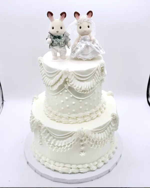 Maria two-tier ivory wedding cake in lambeth piping with swags, ruffles and pearl dots, topped with a pair of toy rabbit figures in a wedding dress and suit, 2026