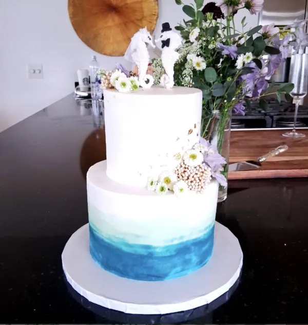 Sunny two-tier wedding cake with a teal watercolor base tier, white daisies and lilac blooms up one side, topped with two white seahorses, 2026