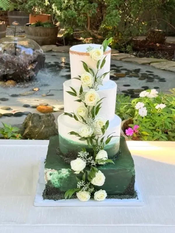 Asia and Max three-tier wedding cake on a square deep green base tier with white roses climbing the white tiers above, beside a garden pond, 2025
