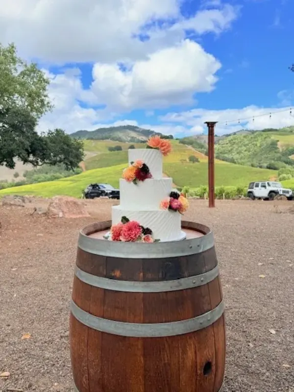 Kacia and Mario three-tier white wedding cake with diagonal combing, coral dahlias and blackberries, on a wine barrel above rolling vineyard hills, 2025