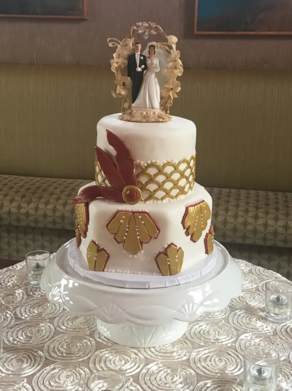 Two-tier white cake with gold and red Art Deco fan motifs and a vintage bride and groom topper