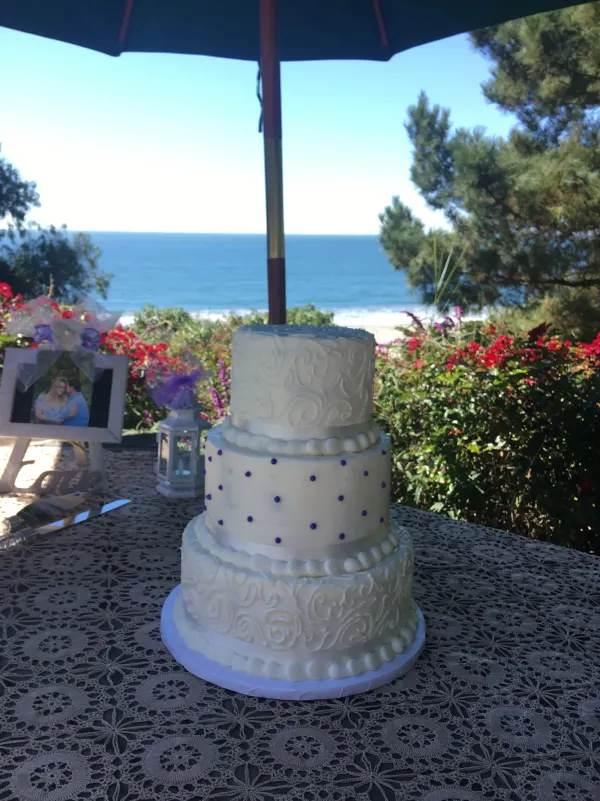 Three-tier white buttercream cake with scroll piping and purple dots on a table above the ocean