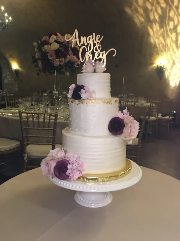 Three-tier white buttercream wedding cake with pink and burgundy flowers and an Angie and Greg topper