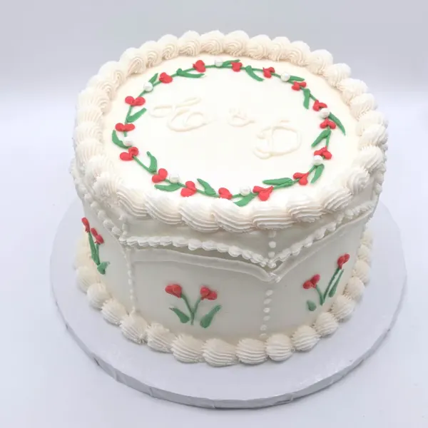 David round ivory wedding cake with a piped red and green berry wreath framing a monogram, shell borders and small piped flowers around the sides, 2025