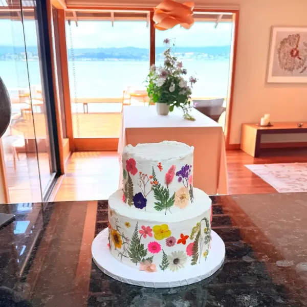 Don two-tier white buttercream wedding cake pressed with edible flowers and ferns, on a stone counter in a room open to the water, 2026
