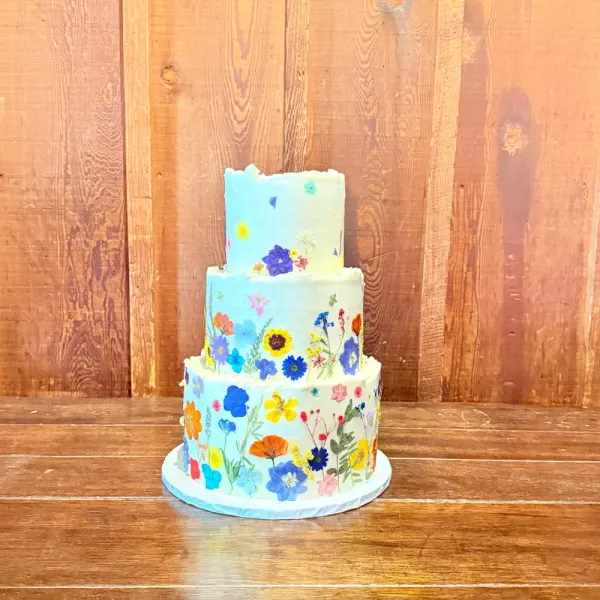 Jiwon three-tier wedding cake washed pale blue and white and covered in pressed edible wildflowers, against a redwood plank wall, 2026