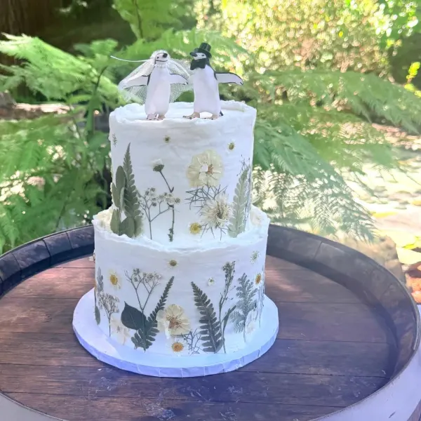 Matt and Ashley two-tier white wedding cake pressed with ferns and dried wildflowers, topped with two penguins in a veil and top hat, on a barrel, 2026