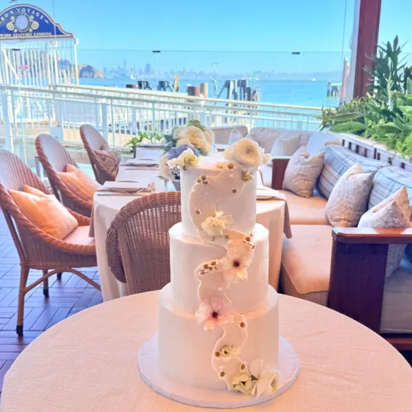 Vanessa three-tier ivory wedding cake with a hand-piped textured ribbon cascading down the front and fresh floral accents, displayed on a round table on an outdoor terrace overlooking the bay, 2026