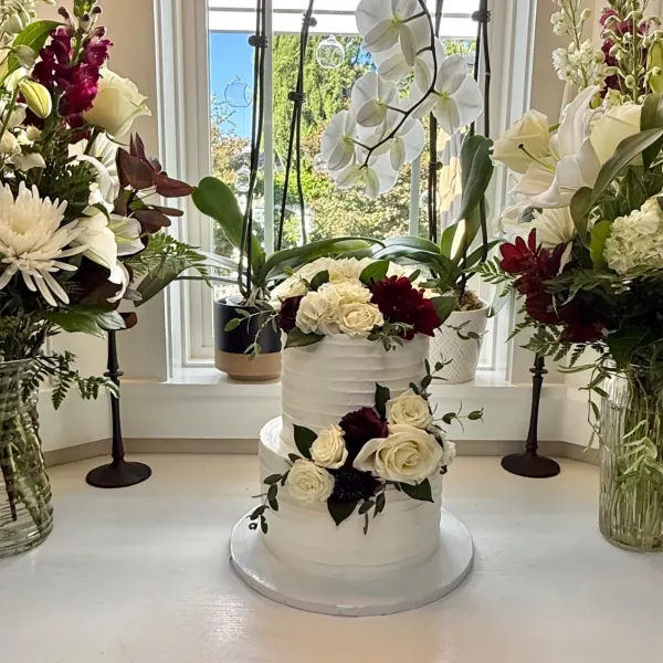 Larissa two tier white buttercream wedding cake with fresh ivory roses and deep burgundy floral accents, displayed on a windowsill surrounded by matching floral arrangements and orchids, 2026