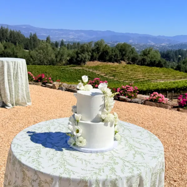 Michaela two-tier white buttercream wedding cake with trailing fresh white roses and baby's breath cascading down one side, displayed on a round table with a vineyard and mountains in the background, 2026