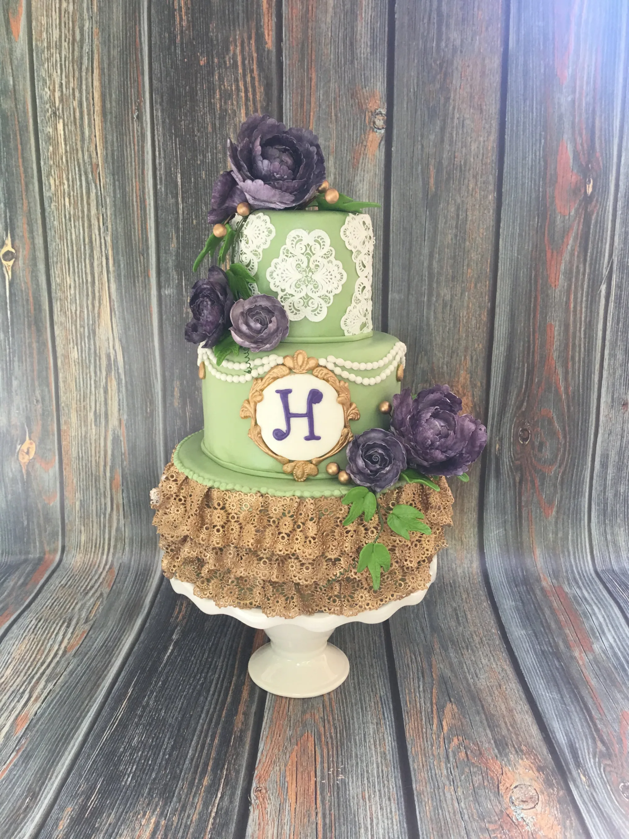 Two-tier sage green wedding cake with a gold lace ruffle base, purple sugar roses and a gold-framed H monogram, 2017