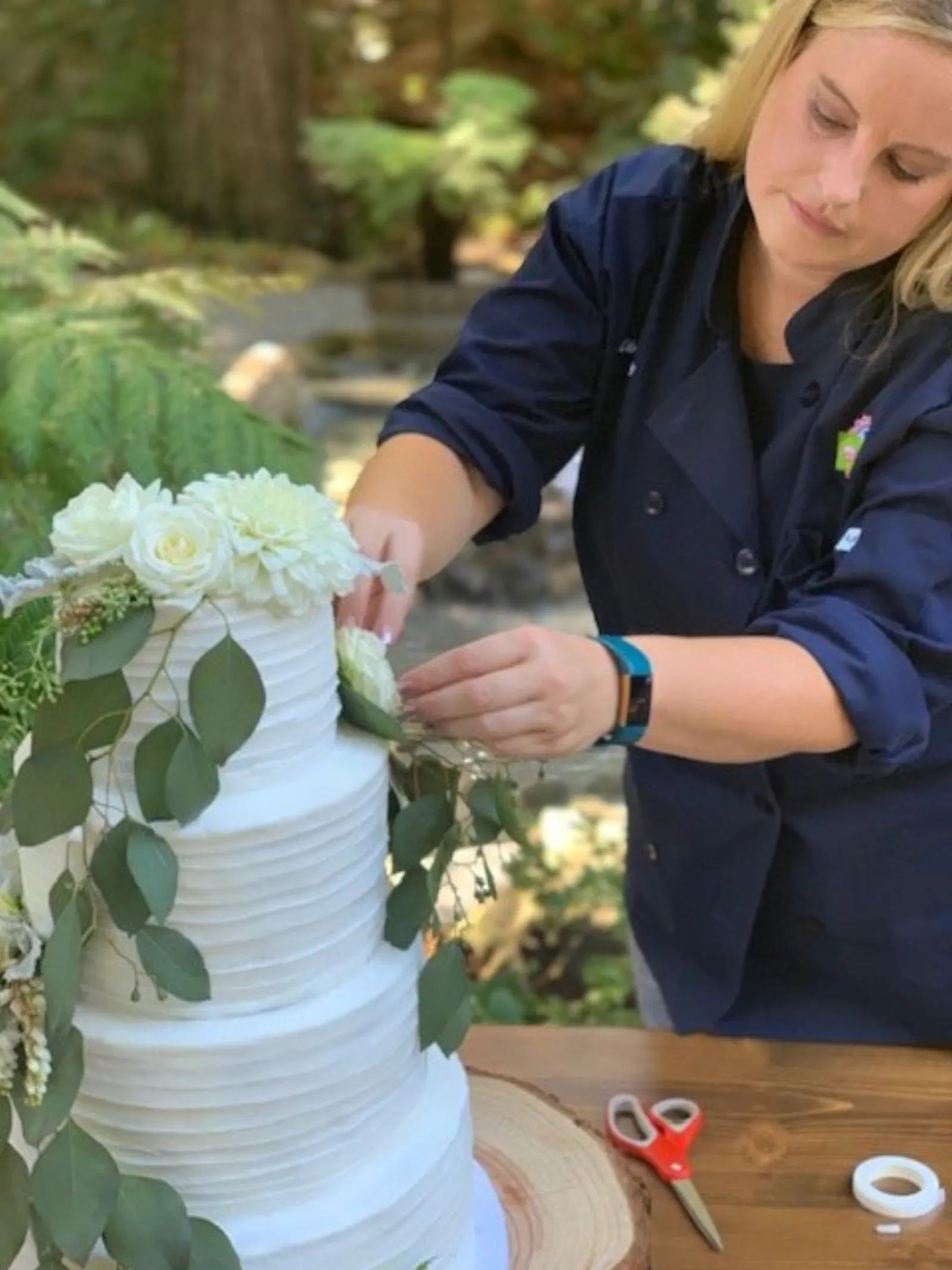 Lindsay Holdorf placing fresh white florals and eucalyptus on a textured buttercream wedding cake at an outdoor redwood venue
